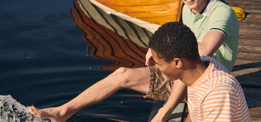 Two men in summer clothing sitting on a dock, one playfully splashing water with his foot, with a wooden boat in the background.