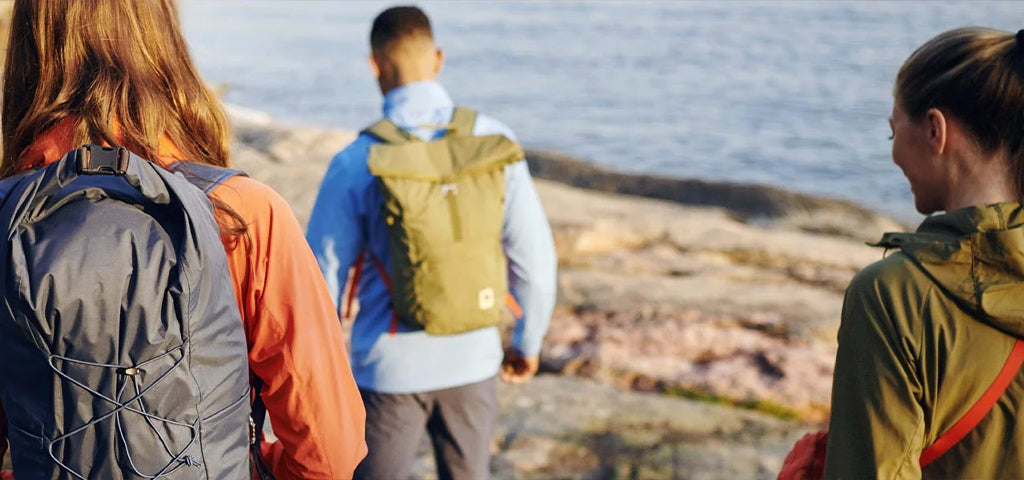 Three people walking along a rocky coastline, wearing backpacks and outdoor apparel, enjoying the serene seaside view.