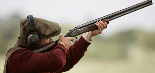 A shooter in traditional fieldwear and hearing protection aiming a Purdey shotgun, with a blurred natural background.