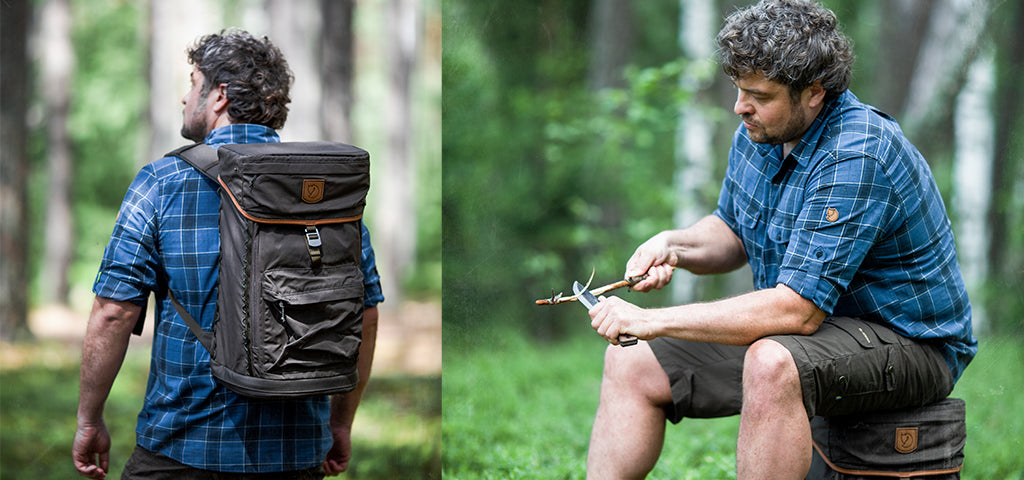 Man wearing a Fjallraven shirt and backpack in the woods, pausing on a hike to carve wood.