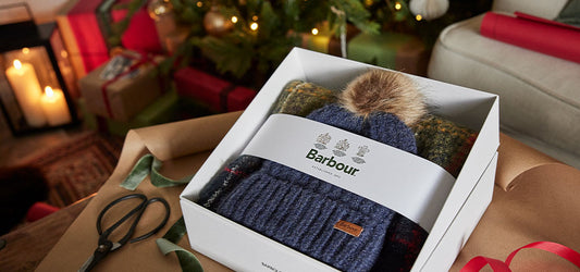 A Barbour gift box with a knitted hat and scarf set, placed on wrapping paper with scissors nearby, in a festive setting with a Christmas tree in the background.