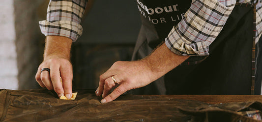 Person in a plaid shirt and Barbour apron applying Barbour wax to a jacket on a workbench.