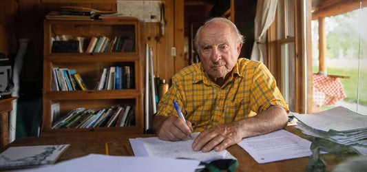 Yvon Chouinard, founder of Patagonia, sitting at a desk with papers, looking at the camera, in a wooden cabin with a bookshelf in the background.