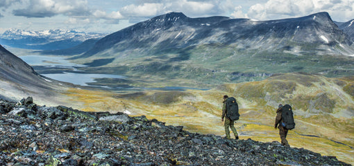 Two hikers with Fjallraven backpacks trekking across a mountainous landscape with a panoramic view of a valley and distant peaks.