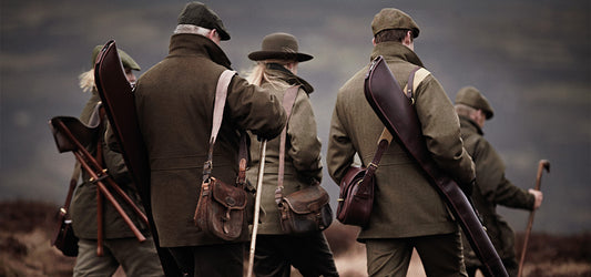 Group of hunters in traditional shooting attire with shotguns and game bags walking through a moorland.