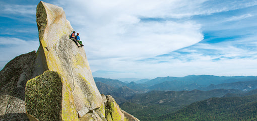 Climbers resting on a cliff edge wearing Patagonia gear with a panoramic view of forested mountains under a clear blue sky.