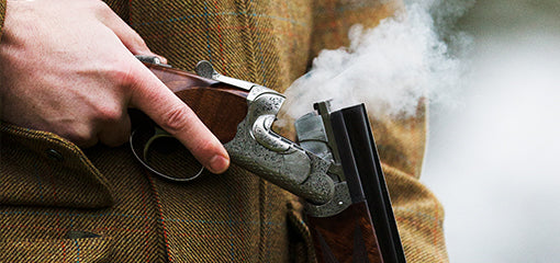 Close-up of a person's hand in a tweed jacket, breaking open a smoking Purdey shotgun after firing.