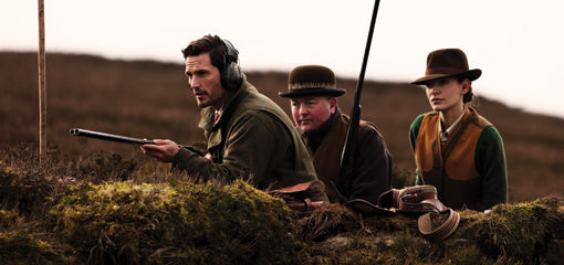 Three hunters lying prone on a moor, with one aiming a shotgun, dressed in outdoor clothing associated with Purdey, in a rural landscape.