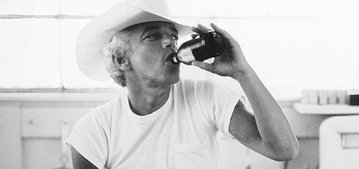 A person in a classic white T-shirt and cowboy hat, associated with RRL clothing by Ralph Lauren, drinking from a bottle.