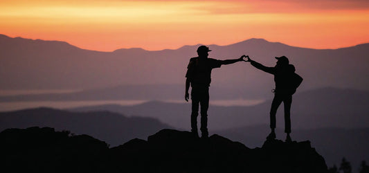 Two people on a mountain peak at sunset forming a heart shape with their hands against an orange sky.