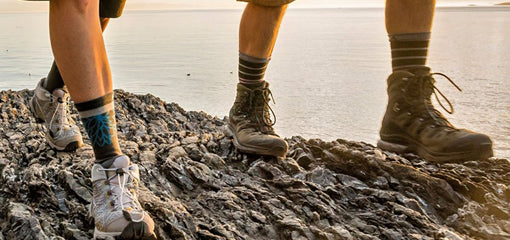 Close-up of two hikers' legs wearing hiking boots and Wigwam socks standing on rocky terrain, with a calm sea in the background.